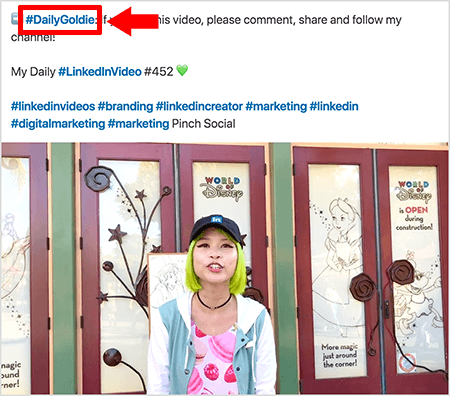 Ez egy képernyőkép, amely bemutatja, hogy Goldie Chan hogyan használ hashtageket a LinkedIn videóbejegyzéseinek szövegében. A piros feliratok a #DailyGoldie hashtagre mutatnak a szövegben, amely egyedülálló a videóbejegyzéseihez, és segít nyomon követni a megosztásokat. A bejegyzés más releváns hashtageket is tartalmaz, amelyek segítenek az embereknek megtalálni a videóját, köztük a #LinkedInVideo. A videoképen Goldie a World of Disney bemutatóján áll néhány ajtó előtt. Zöld hajú ázsiai nő. Fekete LinkedIn sapkát, fekete choker nyakláncot, rózsaszín macaron nyomtatott inget és kék-fehér kabátot visel.